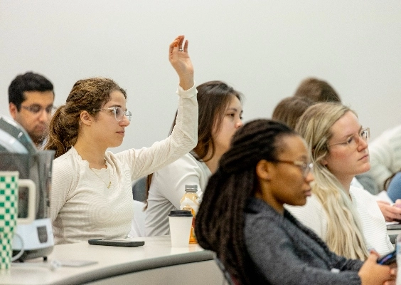 Class sitting at tables. Person in white shirt and glasses raises hand.