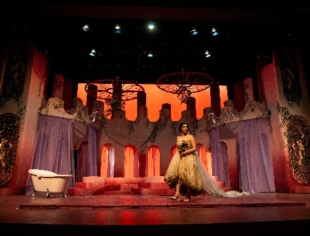 Person in dress on stage with stage decorations behind them including a tub.