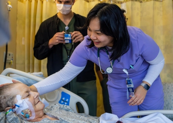 A medical student provides patient care to an older patient in a hospital bed.