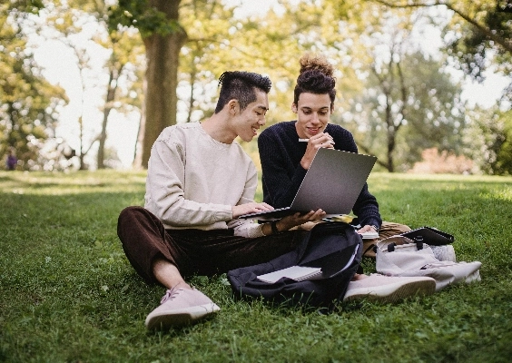 Two graduate students looking at financial aid options on a laptop while sitting outside under some trees.