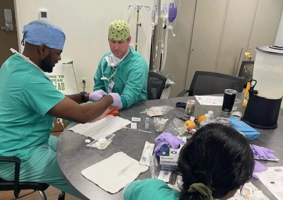 Doctors in scrubs wear gloves and bandanas and sit at table with medical supplies