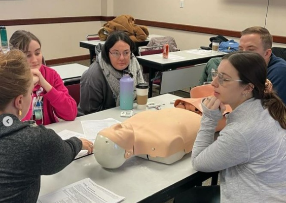 Family medicine residents sit around a table with training dummy on it as they listen to a lecture.