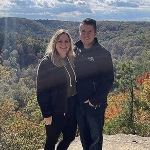 Portrait of Andrea Bastien with her partner standing in front of a forest landscape.