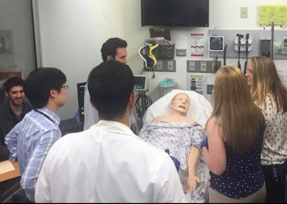 A group of medical students stand around a hospital bed looking on during a simulation training workshop.