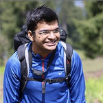 Vasu wearing a blue coat and backpack in a field with trees in the background