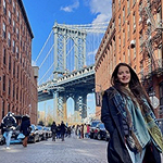 Huda on a city street with buildings and a bridge in the background