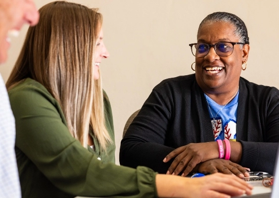 Person wearing glasses and black cardigan smiles at person in green shirt while sitting at a table.