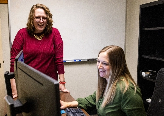 Person in red shirt stands next to person in green shirt sitting at desk behind a computer.