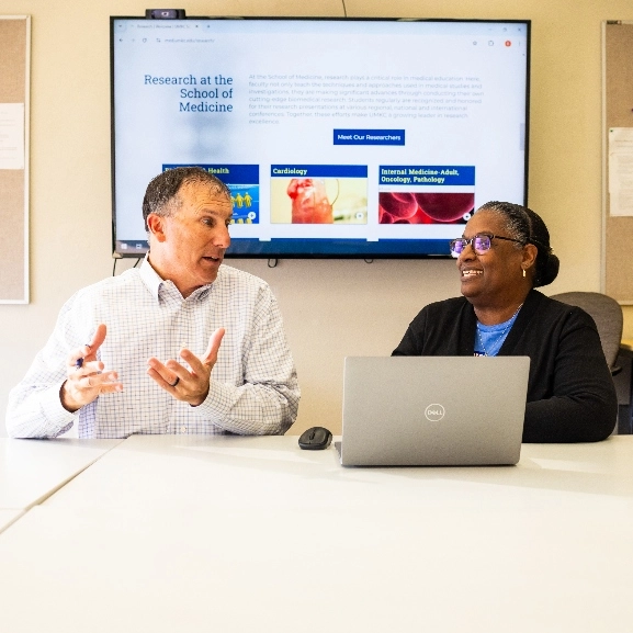 Two people sit at desk talking with screen behind them displaying website. 