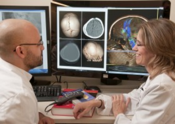 Two people site together with white coats on and look at three computer monitors with scans on them.