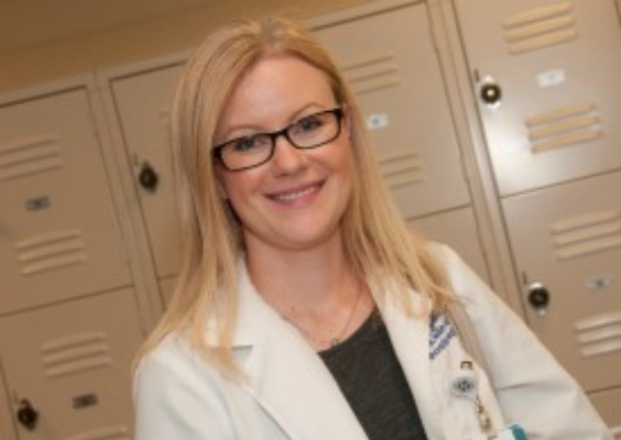 Person smiles at camera wearing white coat and glasses.