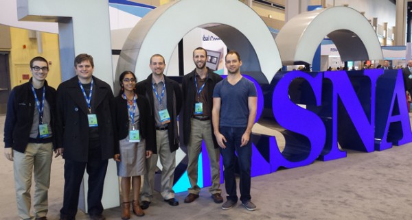 Six students with lanyards and badges stand in front of a lettered sign. All letters are not pictured.