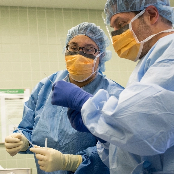 Two doctors in blue scrubs talk with one another while holding handheld surgical tool