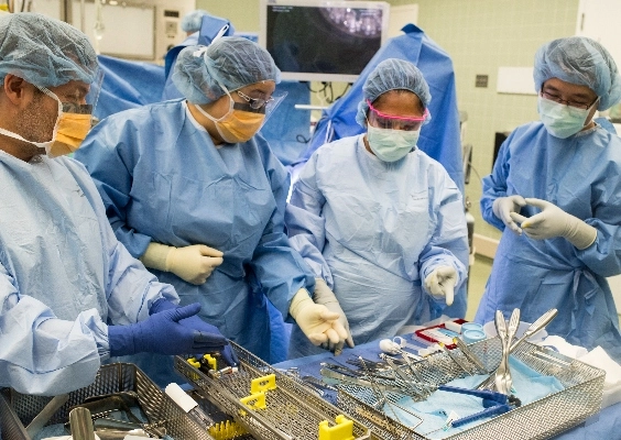 Four doctors wear blue scrubs and stand in front of operating table with equipment