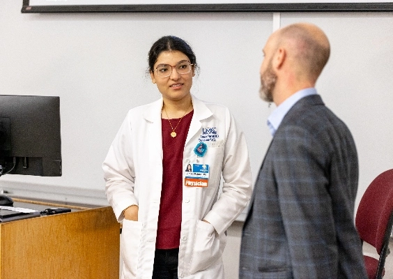 Person wearing physician name tag and white coat talking to someone wearing gray blazer looking away from camera.