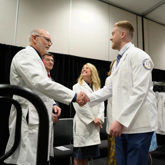 A St. Joseph M.D. program student dressed in his white coat shakes hands with a faculty member. 