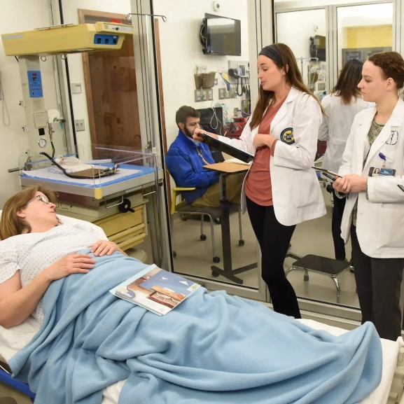 Person with white jacket stands next to person laying down in what appears to be hospital bed